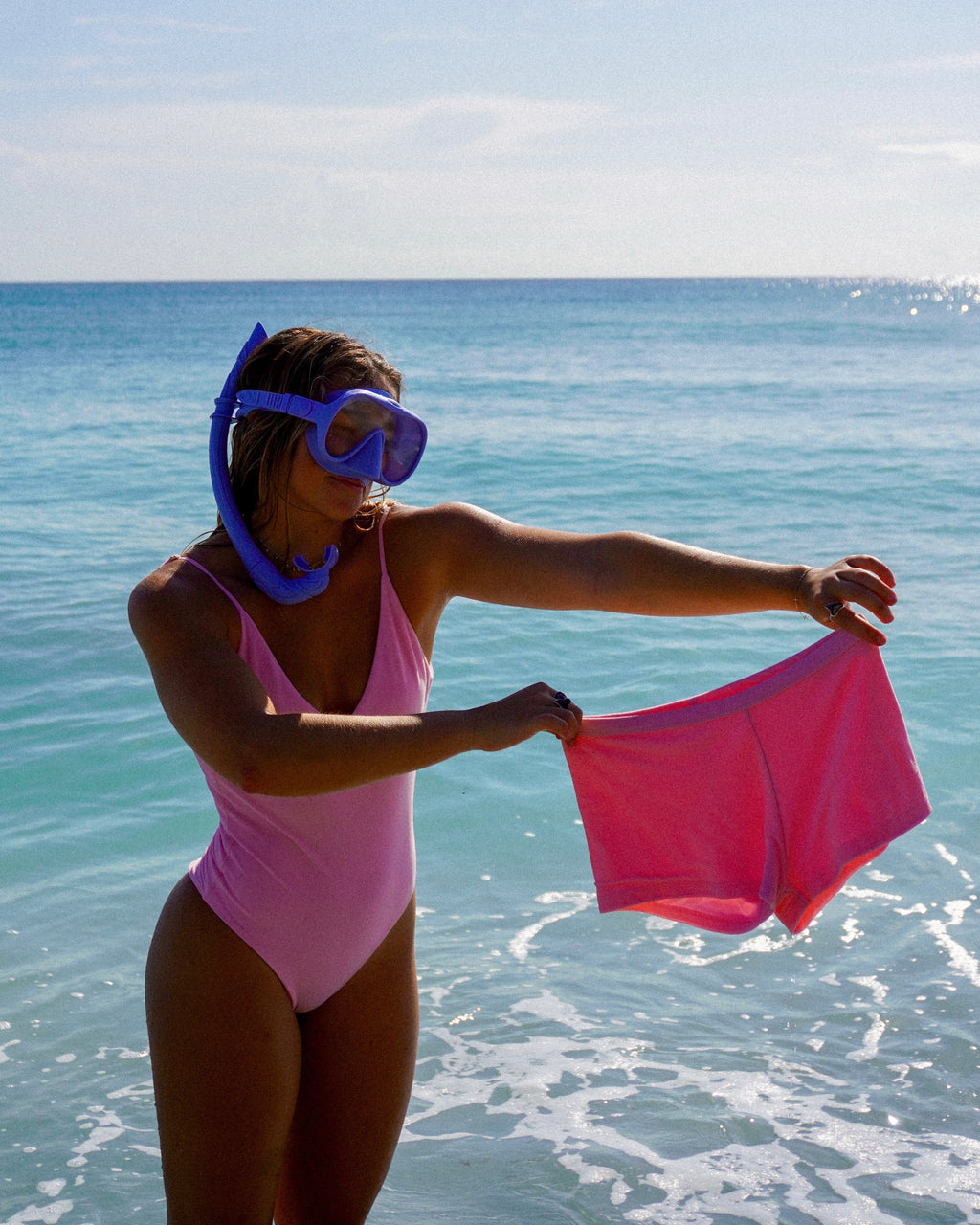Person in pink swimsuit holding pink swim trunks with ocean background