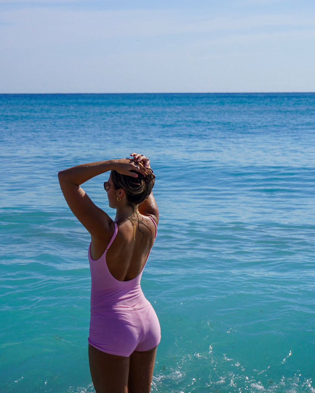 Woman in a pink swimsuit standing by the ocean with clear blue water and sky.