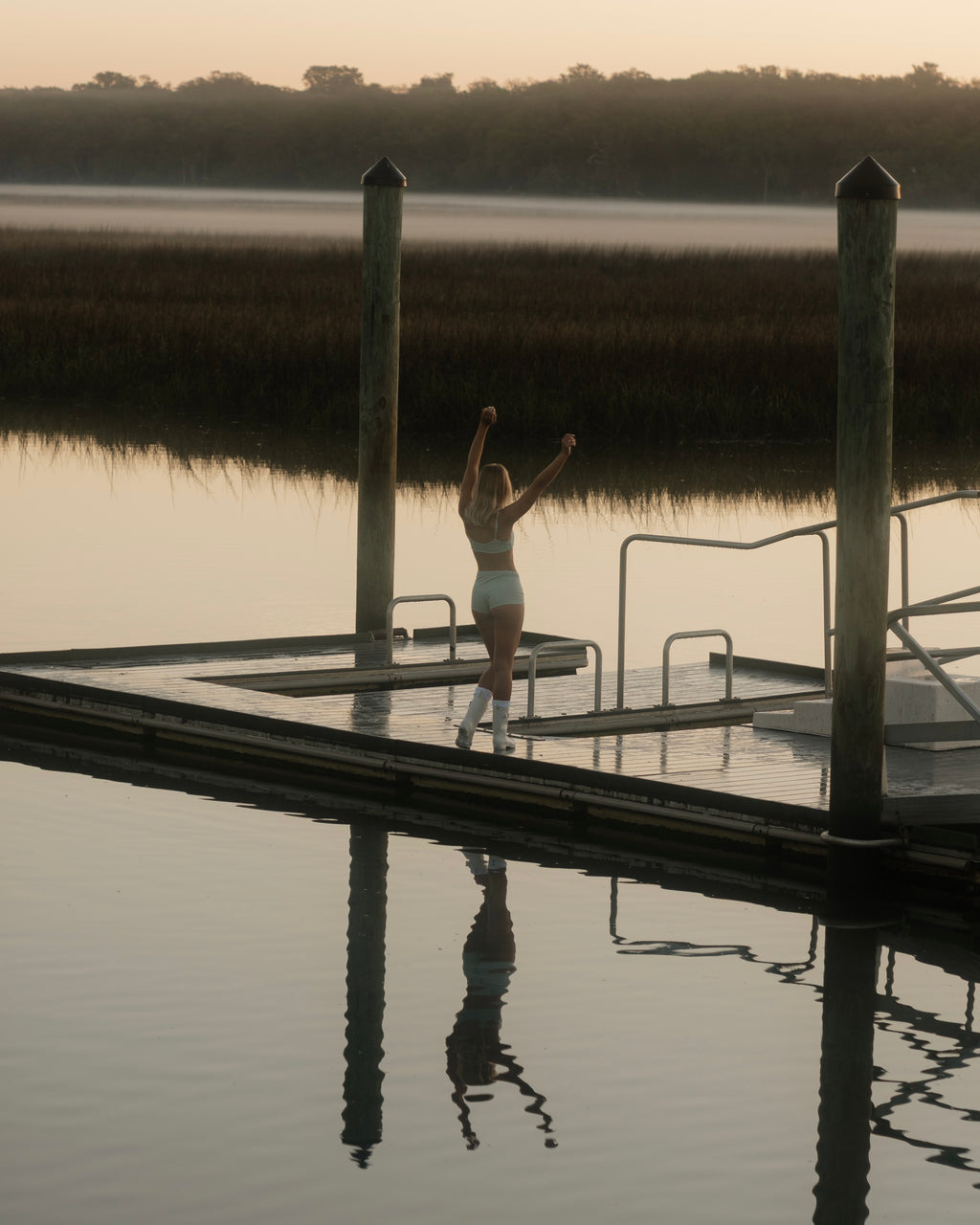 Person standing on a dock by a lake with arms raised at sunset